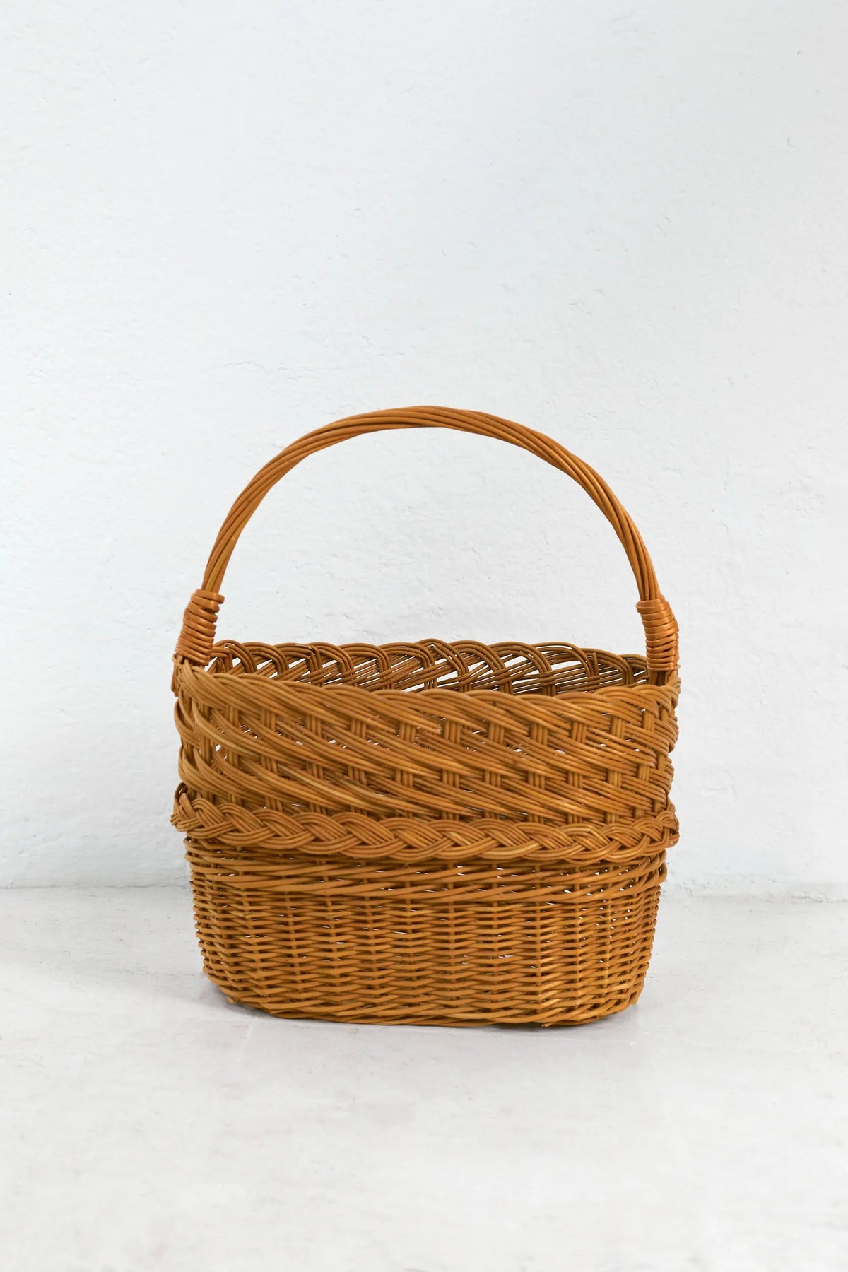 Cesta revistero de mimbre natural con trenzado decorativo, forma ovalada, color marrón y asa superior arqueada.