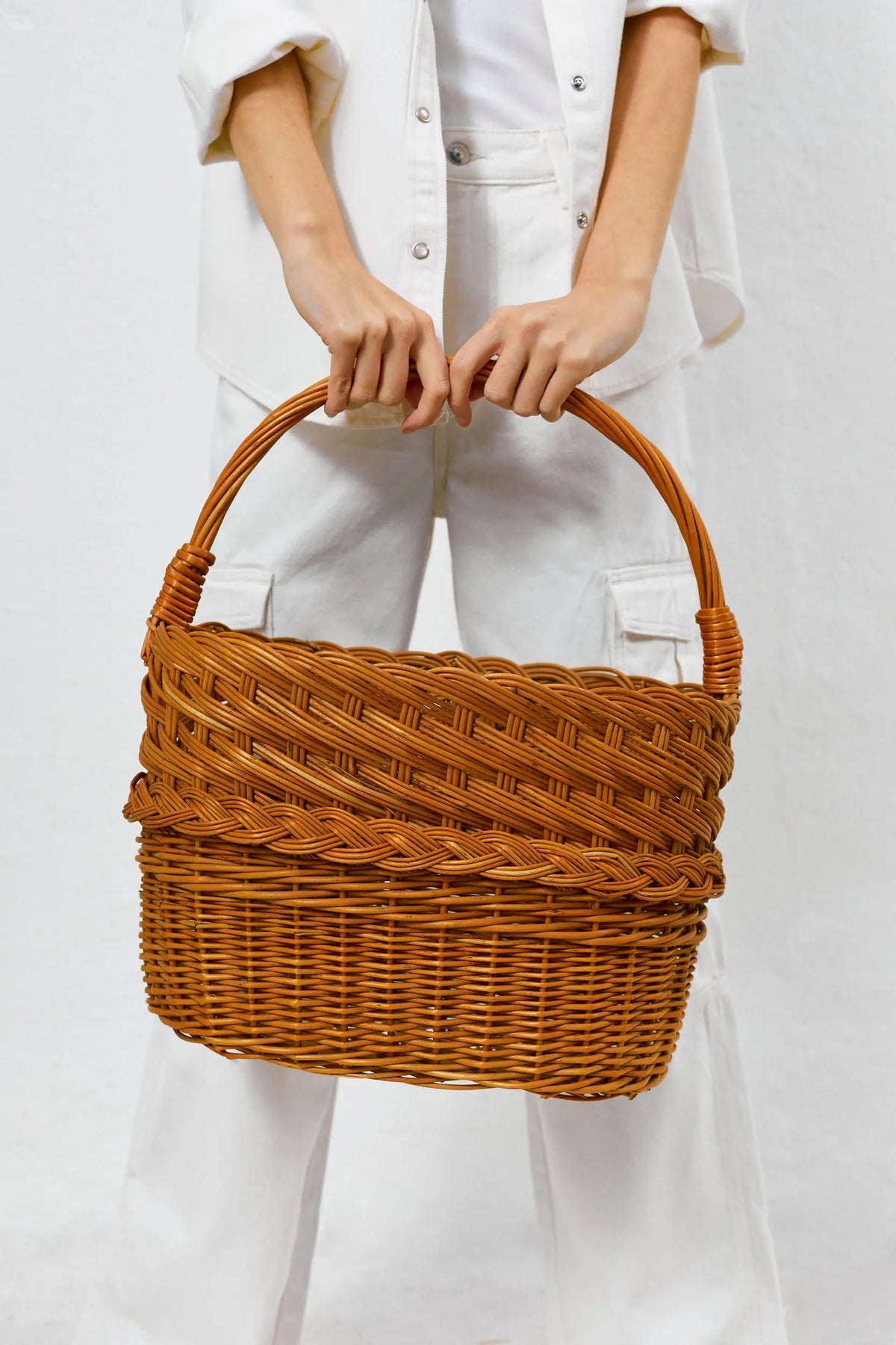 Cesta revistero de mimbre natural con trenzado decorativo y asa arqueada, sostenida por una persona de blanco.