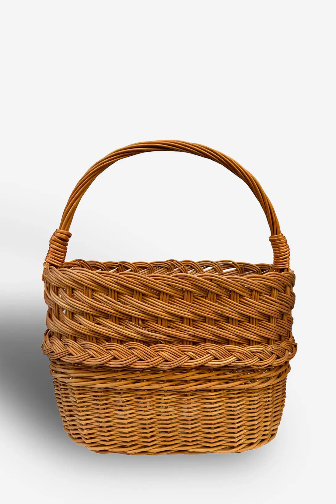 Cesta revistero de mimbre natural con trenzado decorativo, forma ovalada y un asa arqueada, en tono marrón cálido.