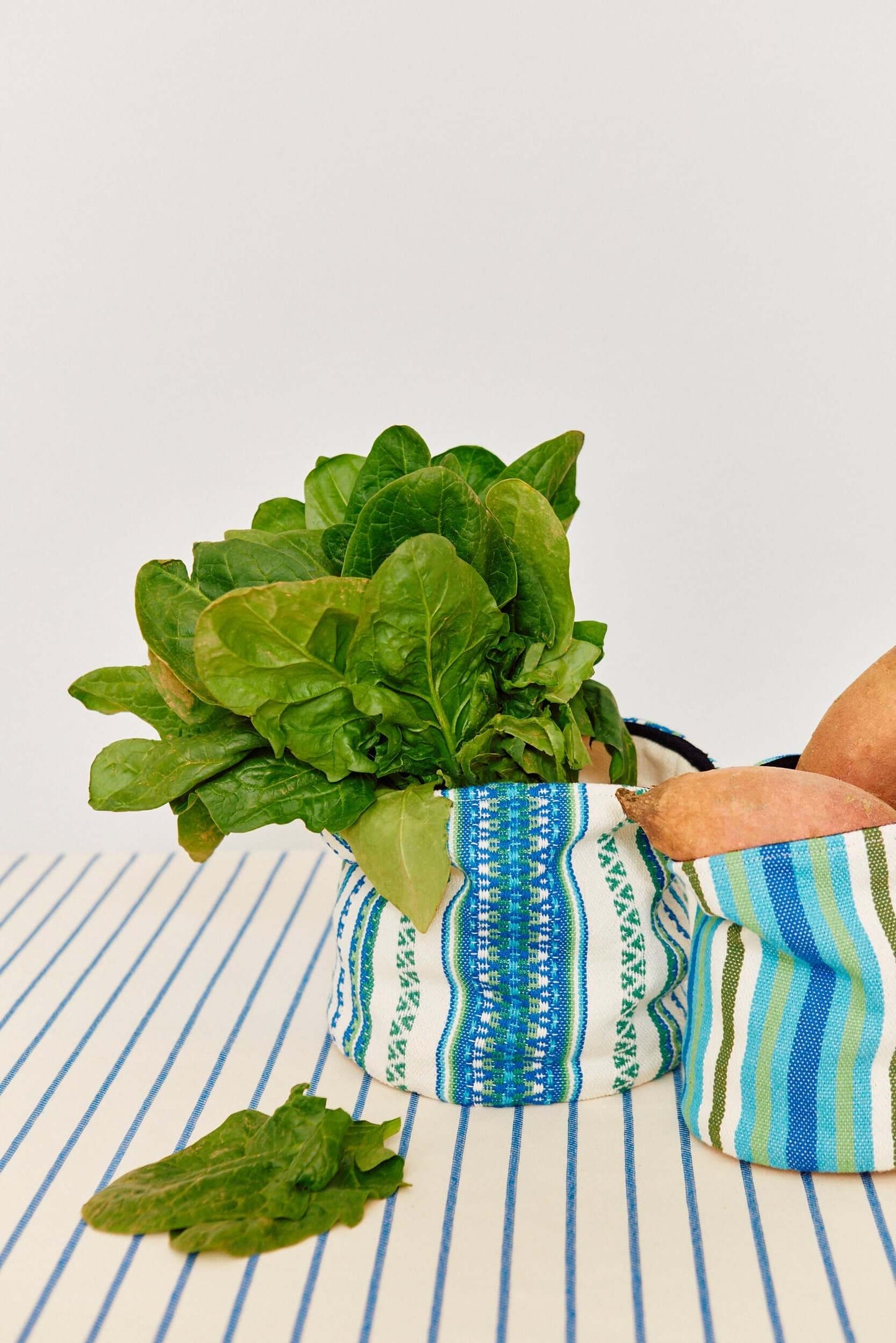 Cestos de tela cilíndricos con rayas blancas, verdes y azules, conteniendo espinacas y camotes sobre una mesa a rayas.