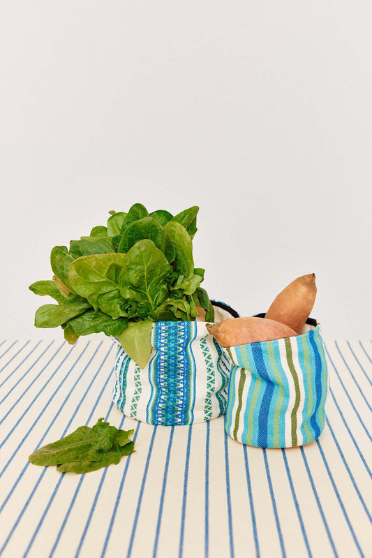 Dos cestos de tela cilíndricos con rayas y patrones geométricos en azul, verde y blanco, con vegetales frescos dentro.