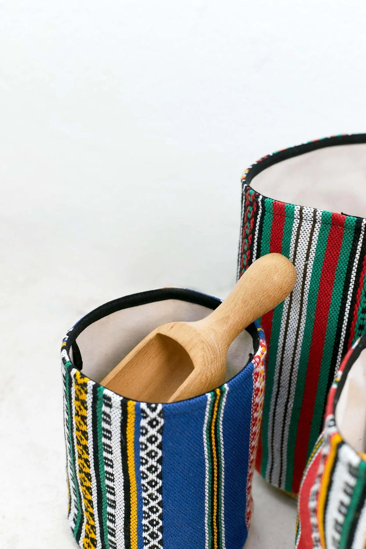Cestos cilíndricos de tela con rayas coloridas y forro interior claro, acompañados por una pala de madera.