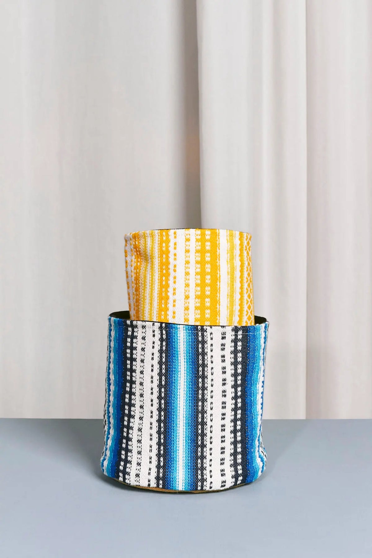 Cesto decorativo de tela estilo alpujarra con rayas verticales en tonos azul, amarillo, blanco y negro sobre fondo neutro.