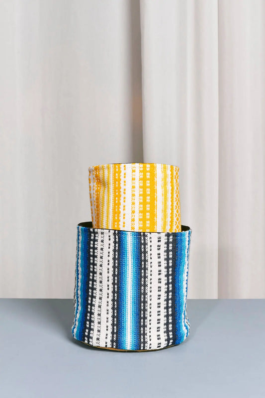 Cesto decorativo de tela estilo alpujarra con rayas verticales en tonos azul, amarillo, blanco y negro sobre fondo neutro.