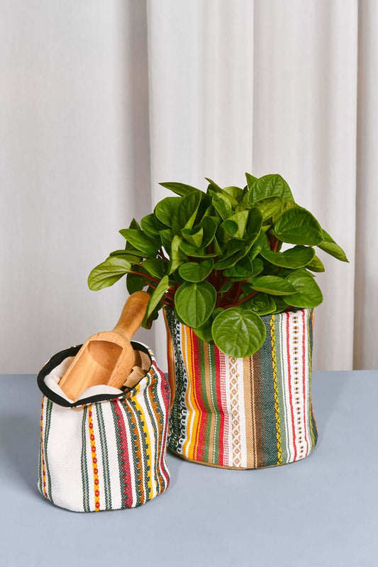 Dos cestos de tela cilíndricos con rayas coloridas; uno contiene una planta verde y el otro una pala de madera.