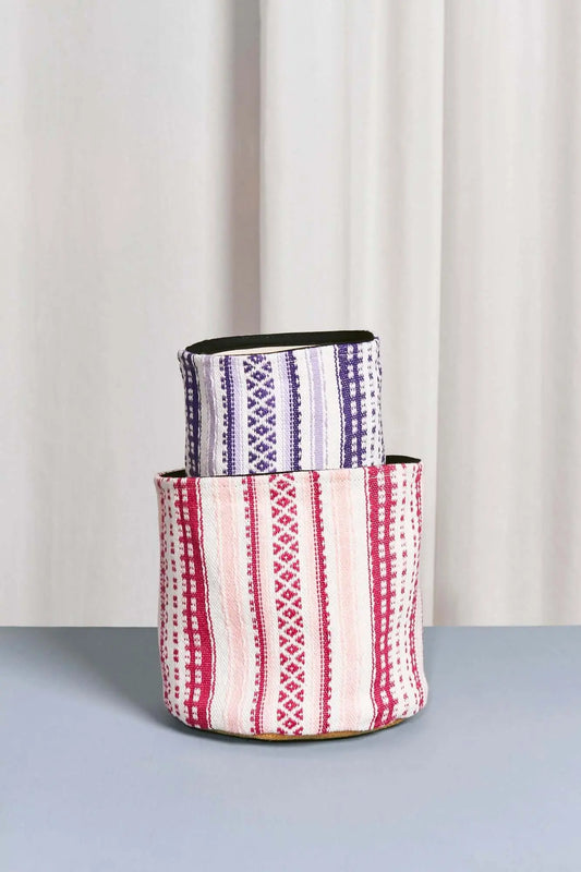Dos cestos textiles cilíndricos, uno sobre otro, con estampado geométrico de rayas en tonos rosa, malva y blanco.
