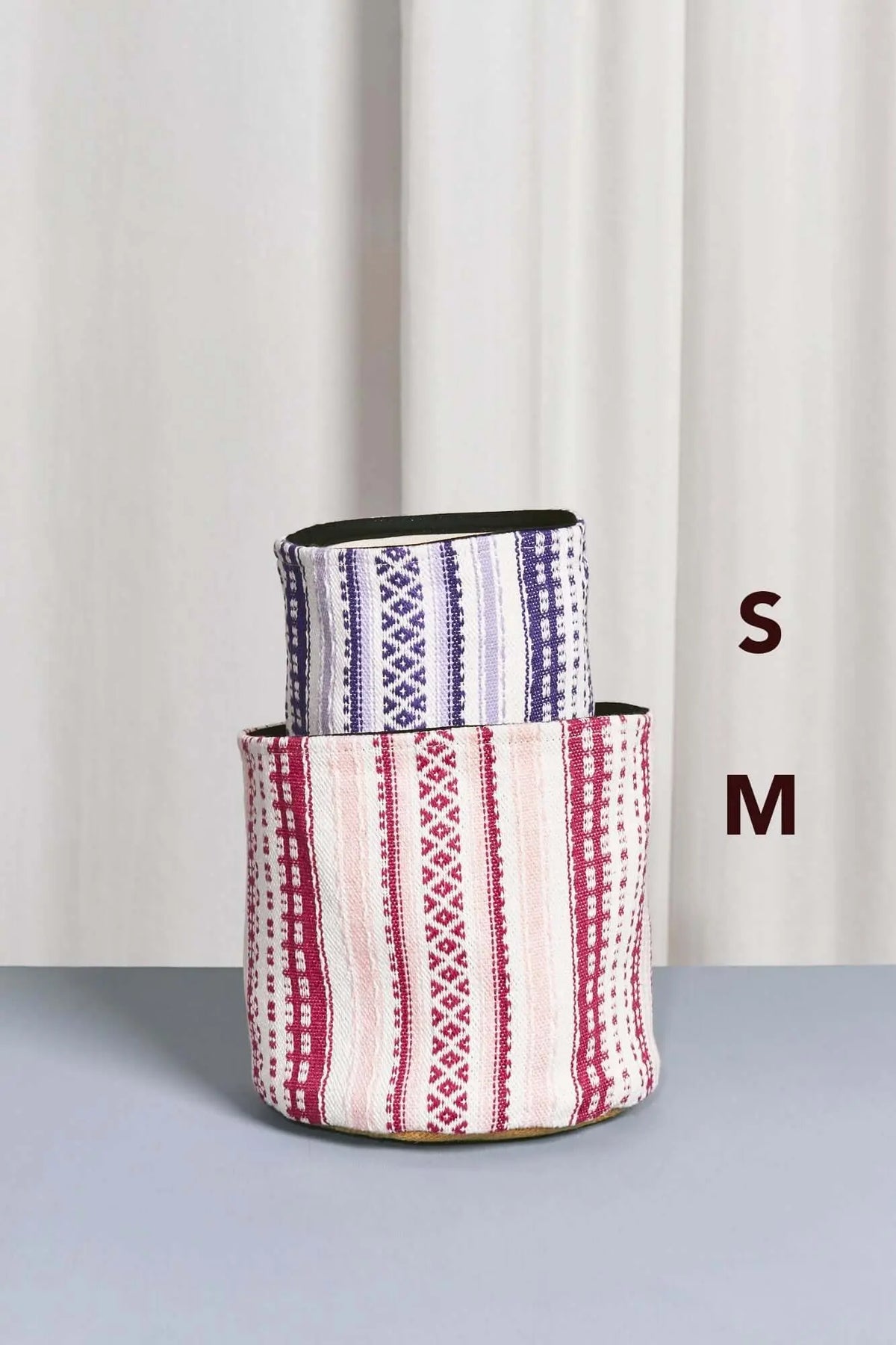 Dos cestos cilíndricos de tela con motivos geométricos y rayas en tonos rosa y malva, uno dentro del otro.