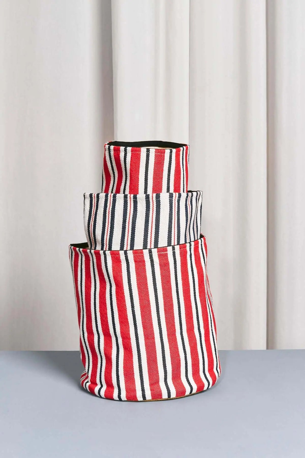 Set de cestos de loneta apilables con estampado de rayas verticales en rojo, azul marino y blanco sobre fondo neutro.