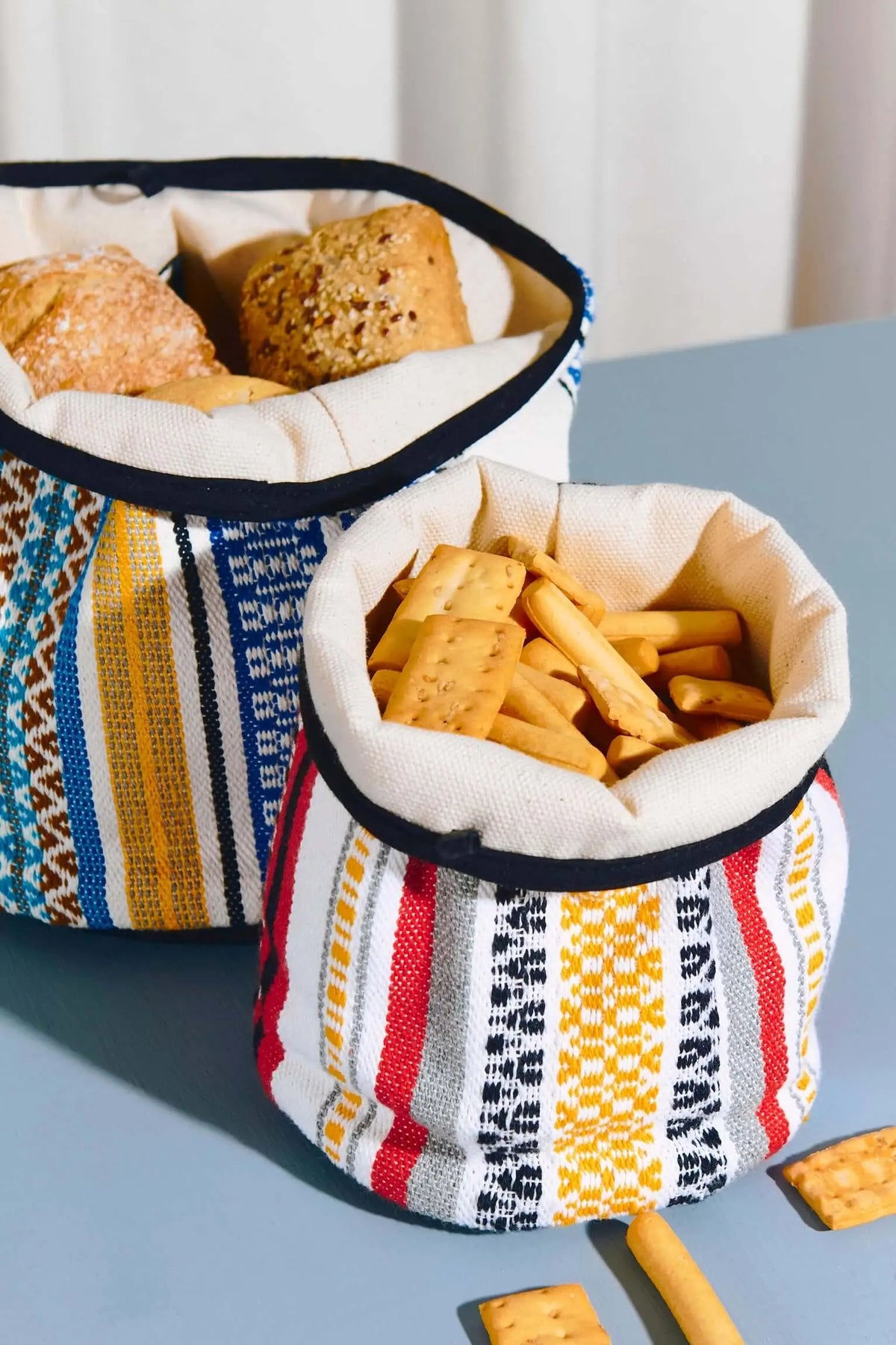 Cestos artesanales de tela con estampado geométrico de rayas en rojo, azul y amarillo, rellenos con pan y galletas.