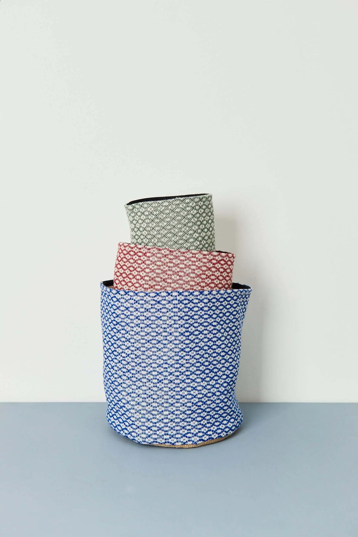 Tres cestos de tela apilados con tejido estampado ojo de perdiz en tonos azul, granate y verde sobre fondo claro.