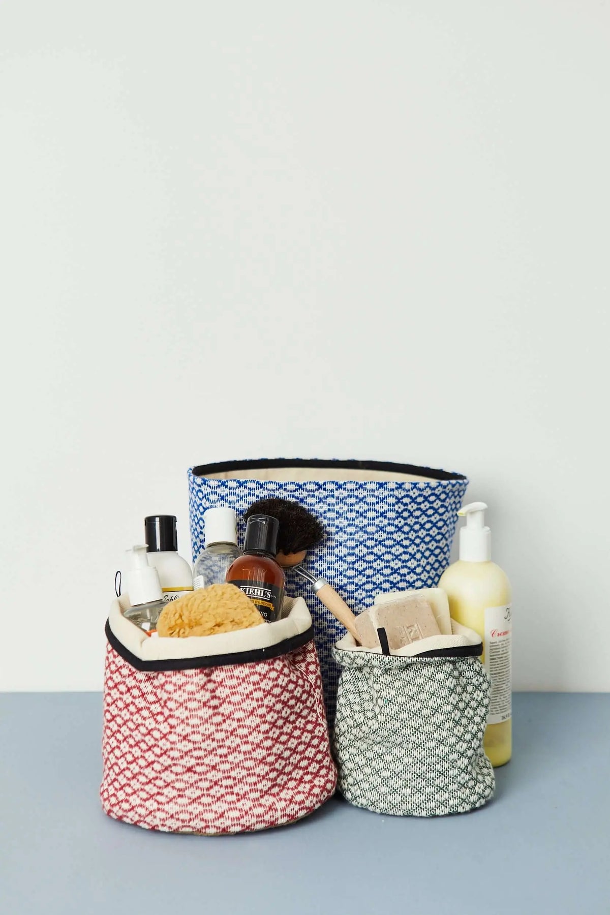Tres cestos de tela con estampado ojo de perdiz en azul, granate y verde, conteniendo productos de aseo personal.