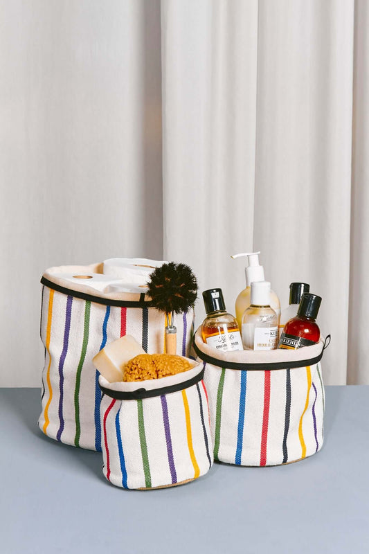 Tres cestos cilíndricos de tela blanca con rayas verticales multicolor y borde negro, llenos de artículos de aseo.