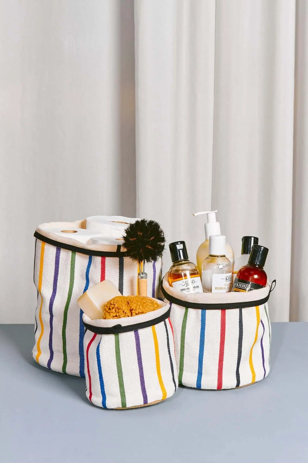 Tres cestos cilíndricos de tela blanca con rayas verticales multicolor y borde negro, llenos de artículos de aseo.