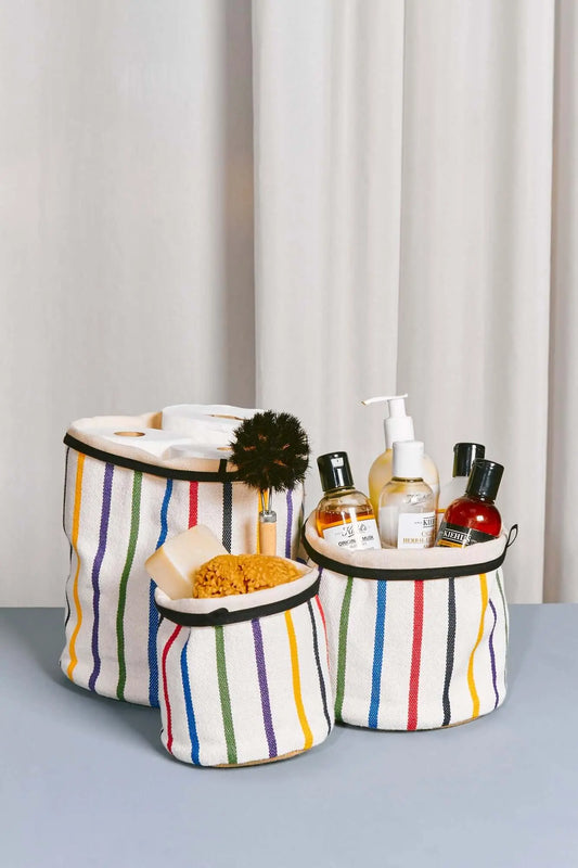 Tres cestos cilíndricos de tela blanca con rayas verticales multicolor y borde negro, llenos de artículos de aseo.