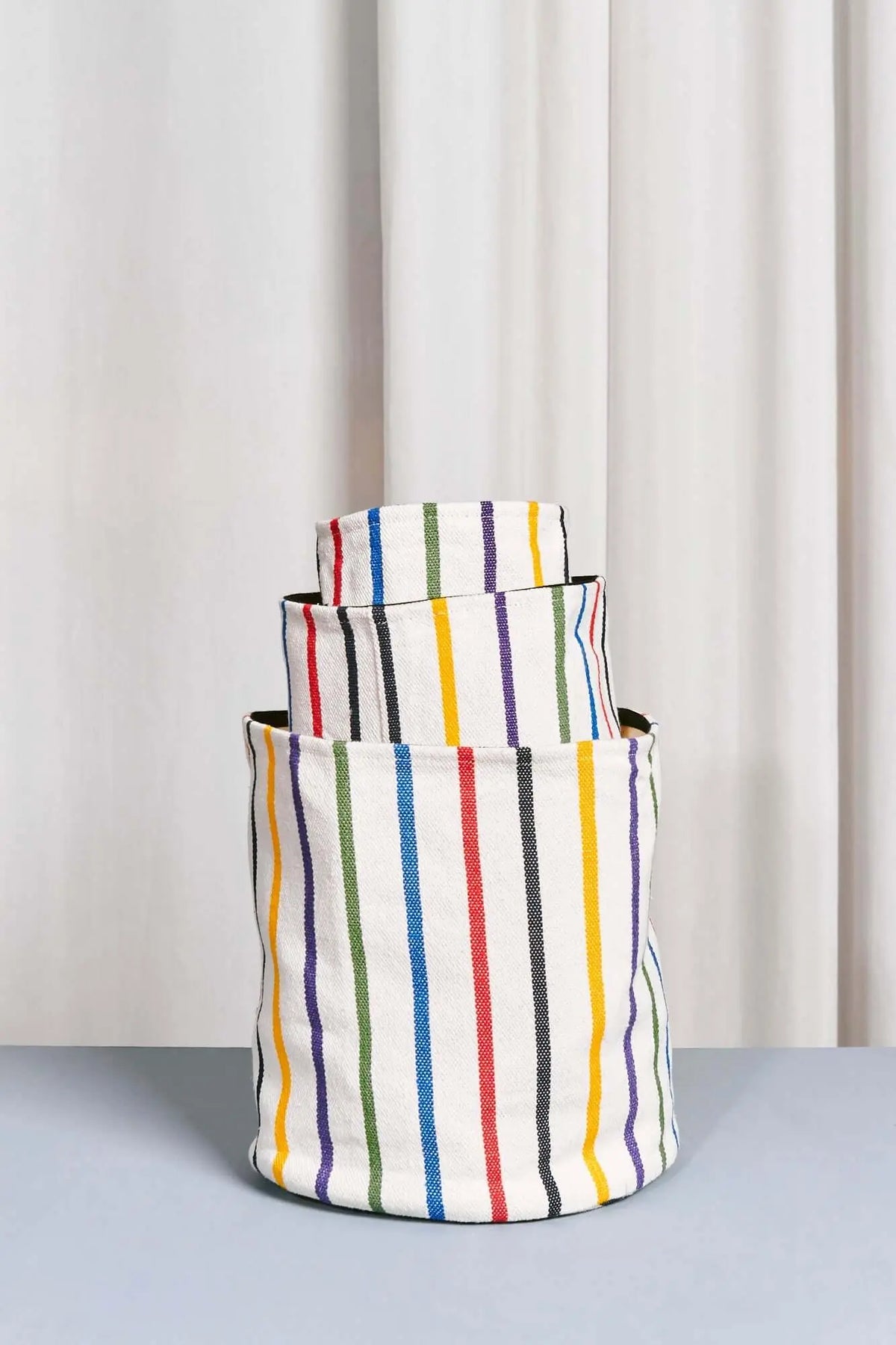 Tres cestos textiles cilíndricos apilados, de fondo blanco con rayas verticales coloridas y estilo artesanal.