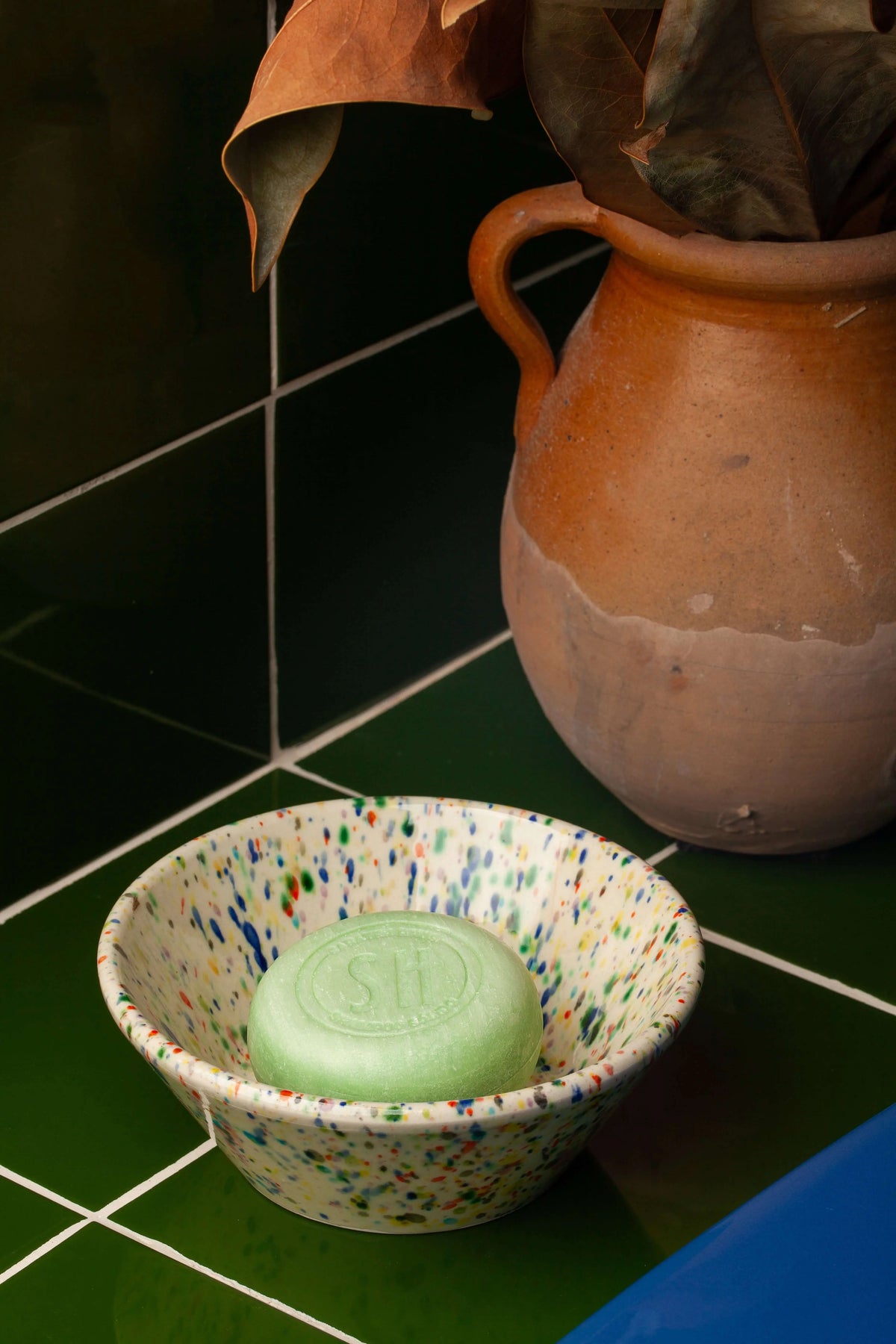 Round light green solid shampoo bar in a speckled bowl, set against dark green tiles and a rustic clay pitcher.