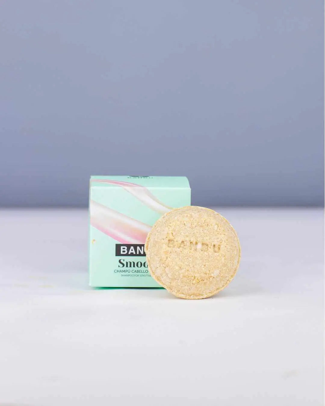 Champú sólido Banbu Smooth con textura granulada frente a su caja de cartón verde claro sobre fondo gris azulado.