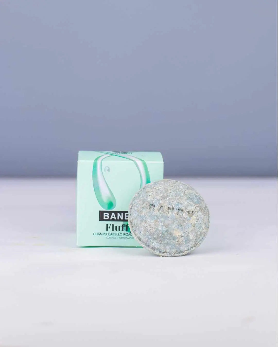 Champú sólido redondo grisáceo frente a su caja de cartón verde claro de la marca Banbu para cabello rizado.