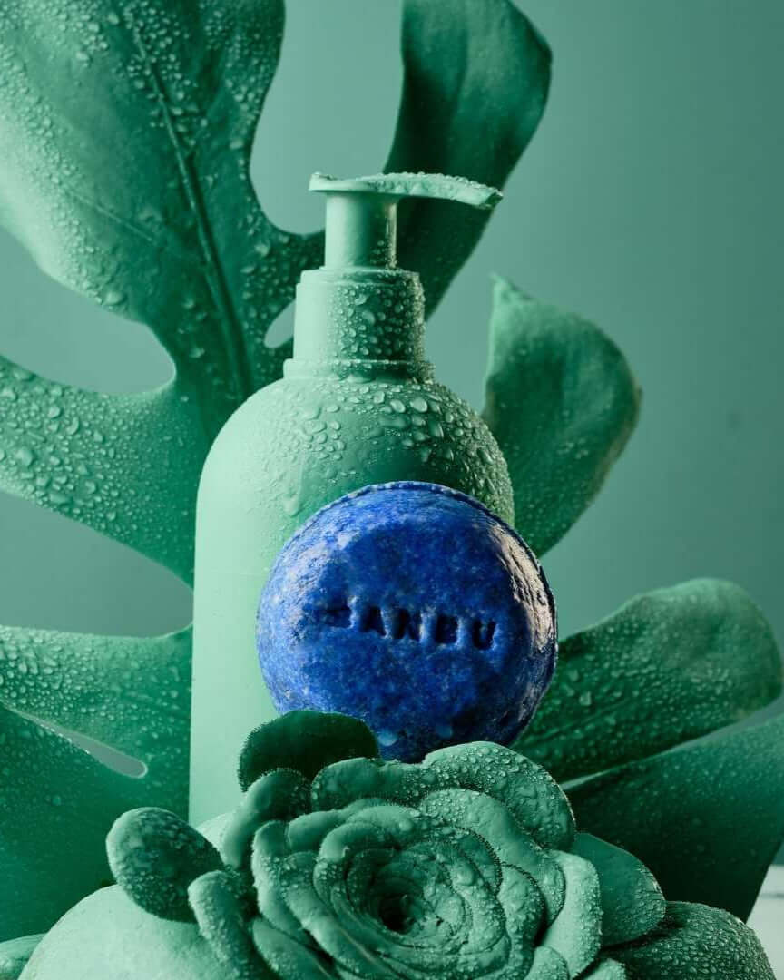 Champú sólido azul Banbu y botella verde entre hojas húmedas con gotas de agua sobre fondo verde.