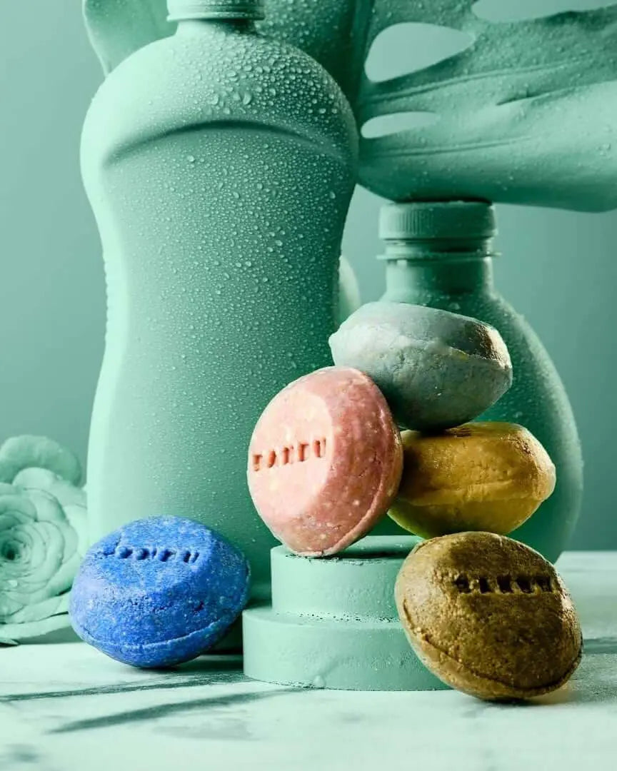 Champú sólido Banbu de colores sobre pedestal verde, con envases de plástico húmedos de fondo, estilo ecológico.