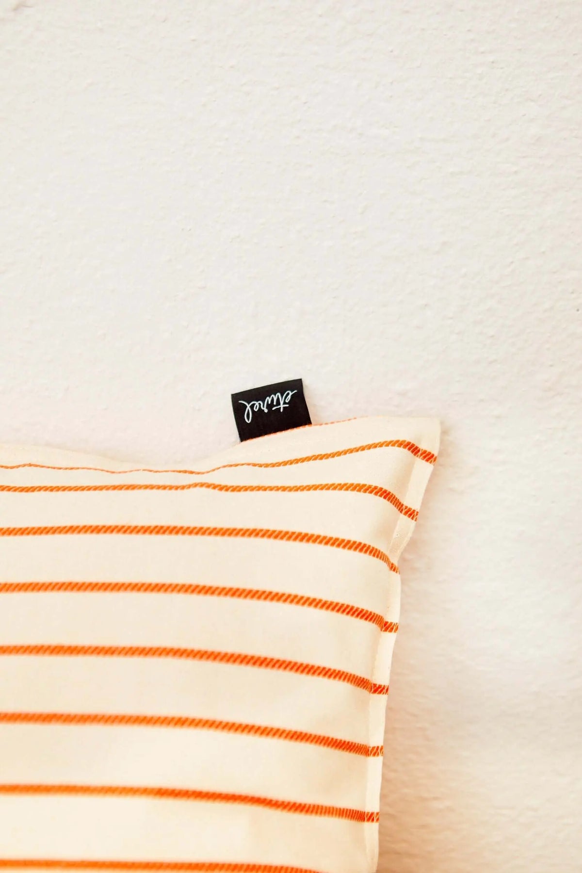 Rectangular off-white cushion cover with horizontal orange striped texture and a small black brand label.