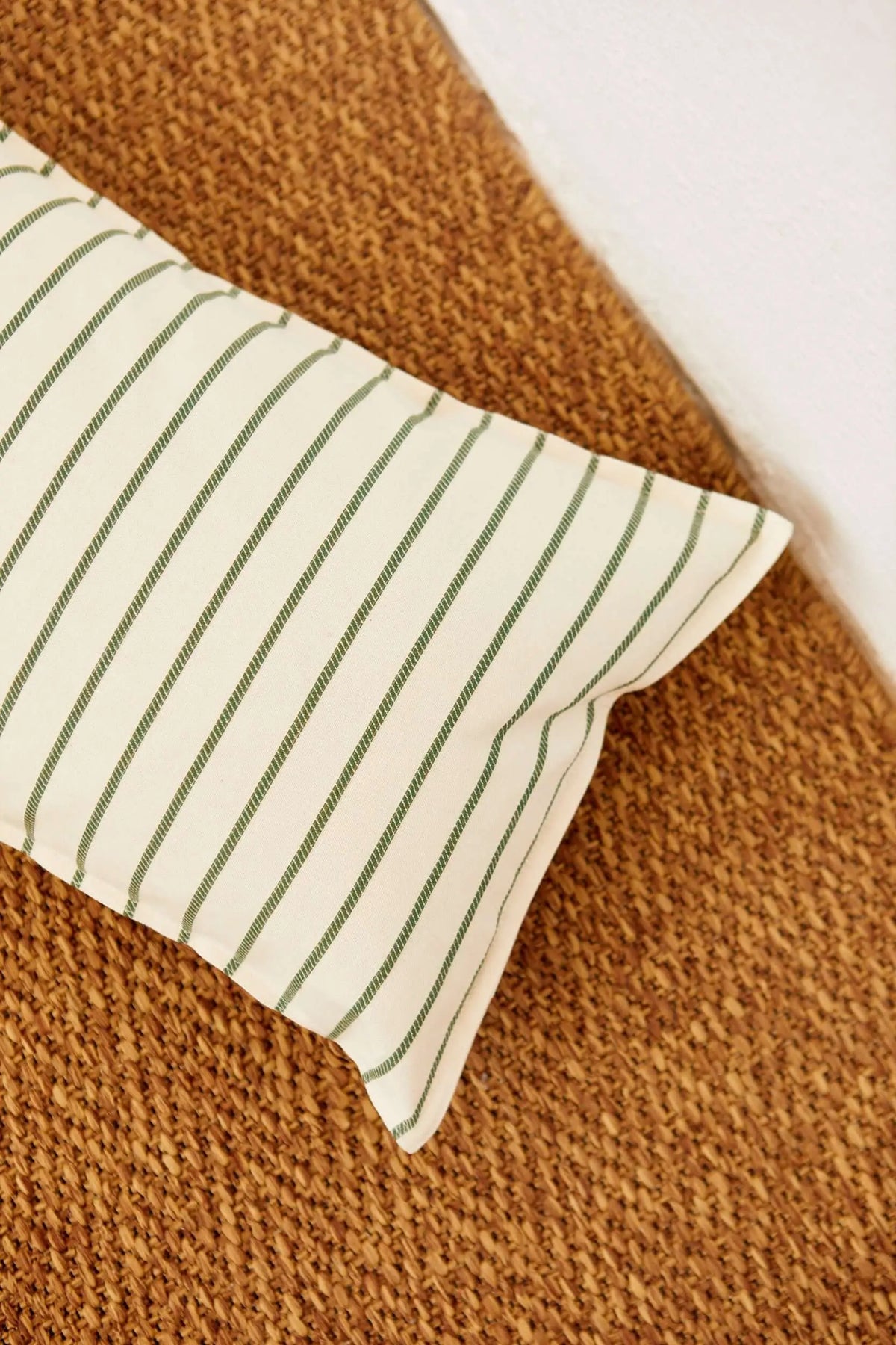 Rectangular off-white cushion cover with thin green stripes, resting on a textured brown woven surface.