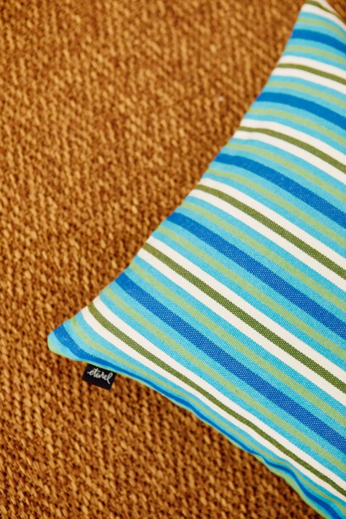 Funda de cojín cuadrada con estampado de rayas horizontales en tonos azules, verdes y blancos sobre fondo de mimbre.