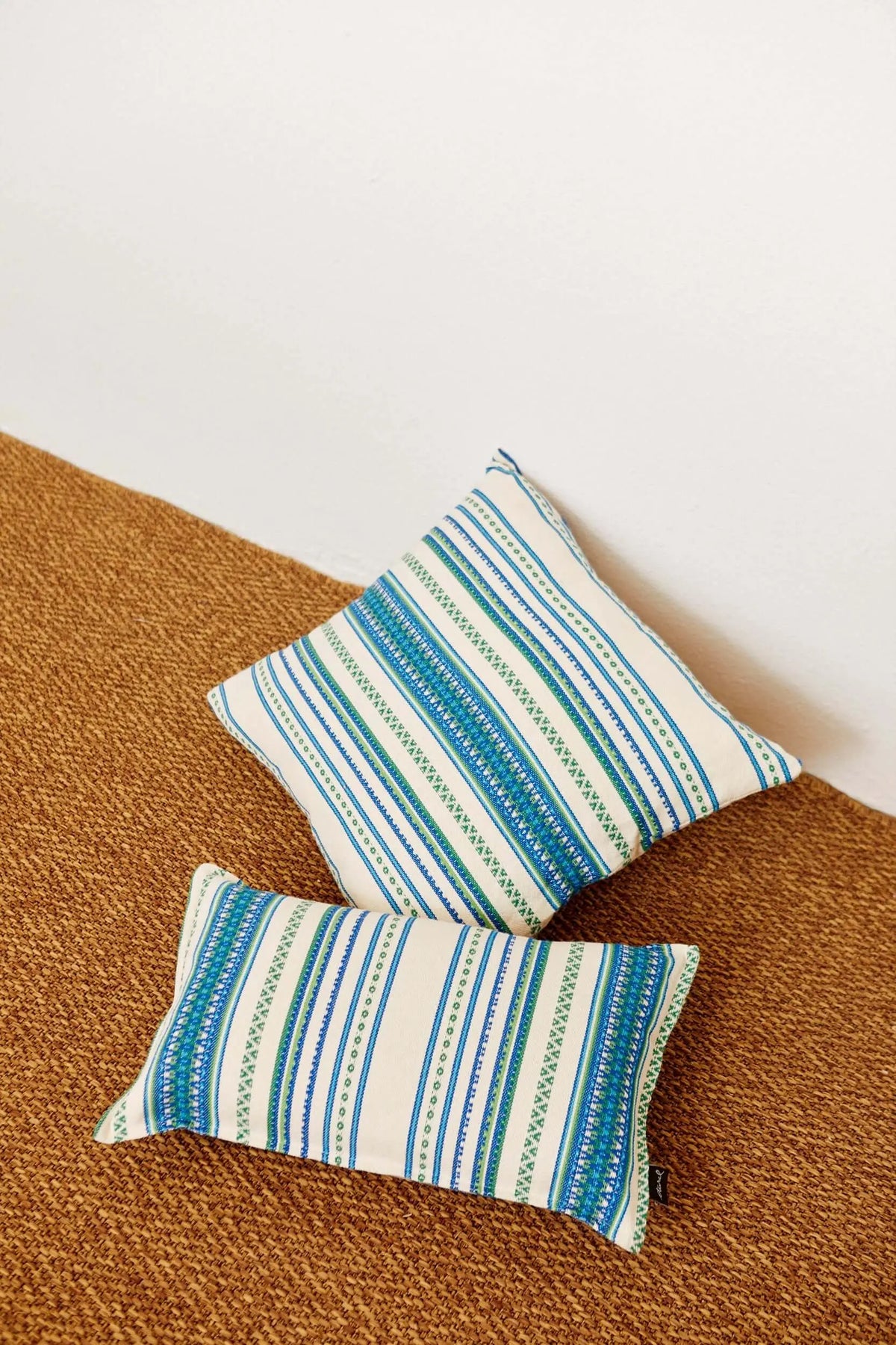 Dos fundas de cojín de tejido manchego con rayas geométricas en tonos azul y verde sobre fondo crema, estilo artesanal.