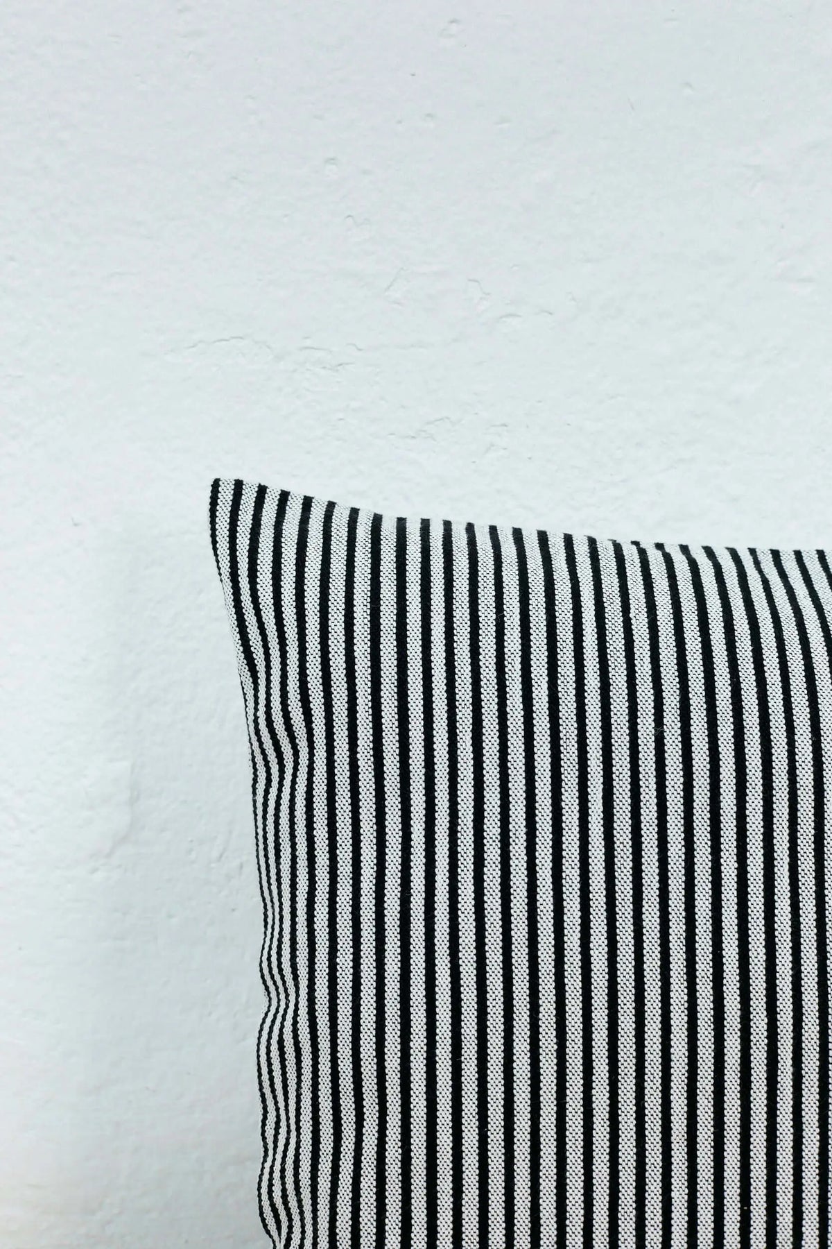 Funda de cojín cuadrada de loneta con patrón de rayas verticales finas en blanco y negro sobre fondo de pared blanca.