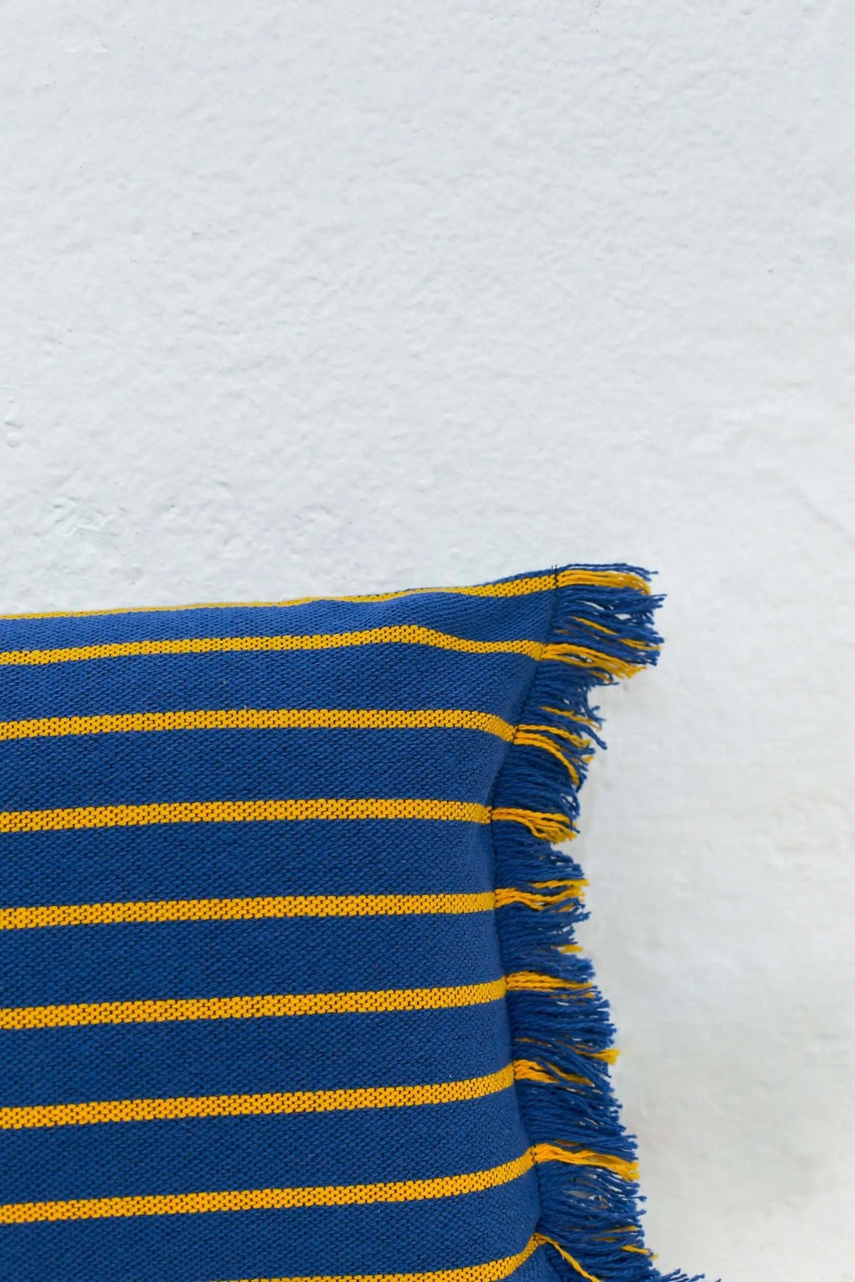 Rectangular blue cushion cover with thin yellow horizontal stripes and fringed edges against a white textured wall.