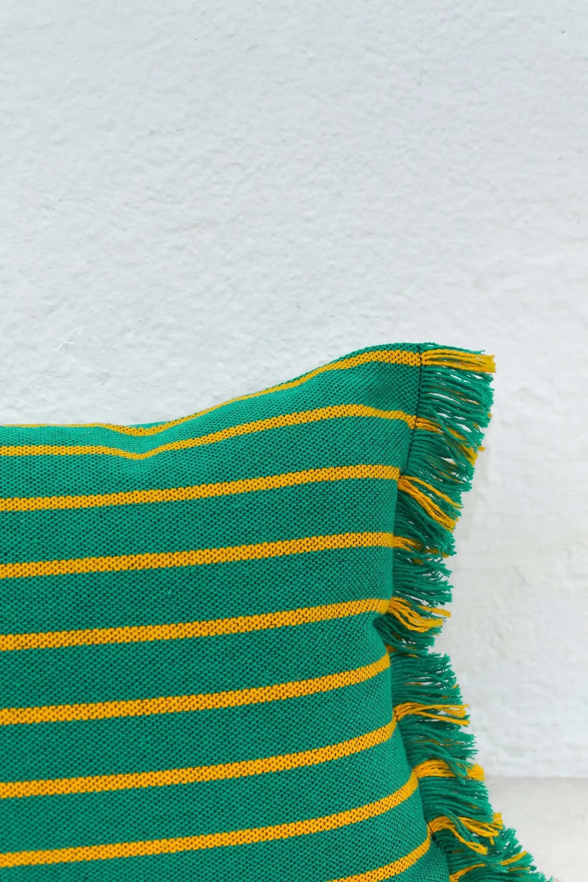 Funda de cojín rectangular verde y amarilla a rayas con flecos decorativos en un lateral sobre fondo blanco.