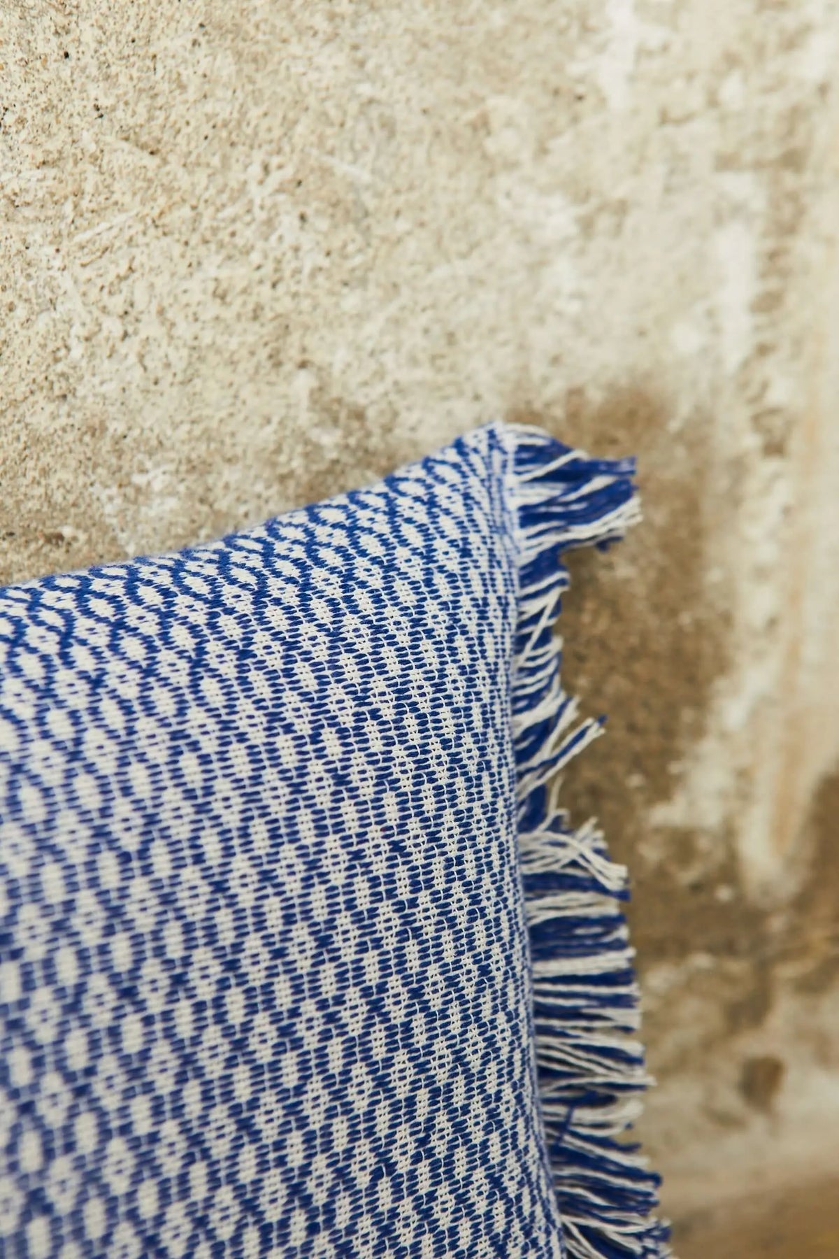 Funda de cojín azul con estampado geométrico de ojo de perdiz y bordes con flecos, sobre fondo de pared texturizada.