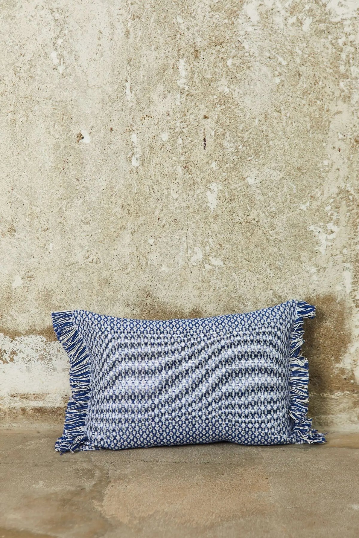 Rectangular blue and white patterned cushion cover with fringe edges, set against a rustic textured stone wall.