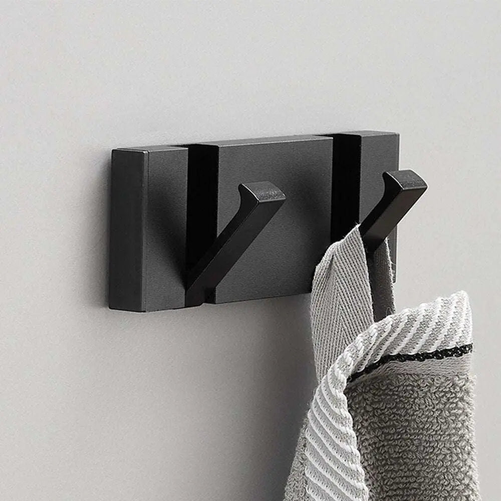 Perchero de pared moderno de metal negro mate con dos ganchos, sosteniendo una toalla gris sobre pared clara.