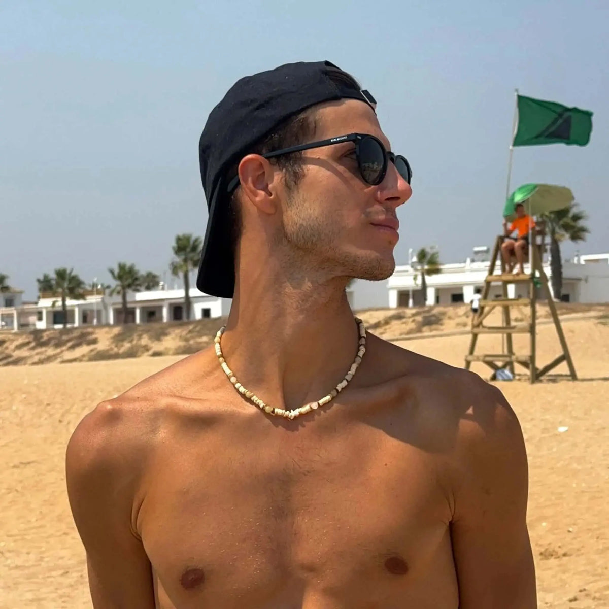 Hombre con gafas de sol de montura negra y lentes polarizadas verdes en la playa, luciendo un estilo casual de verano.