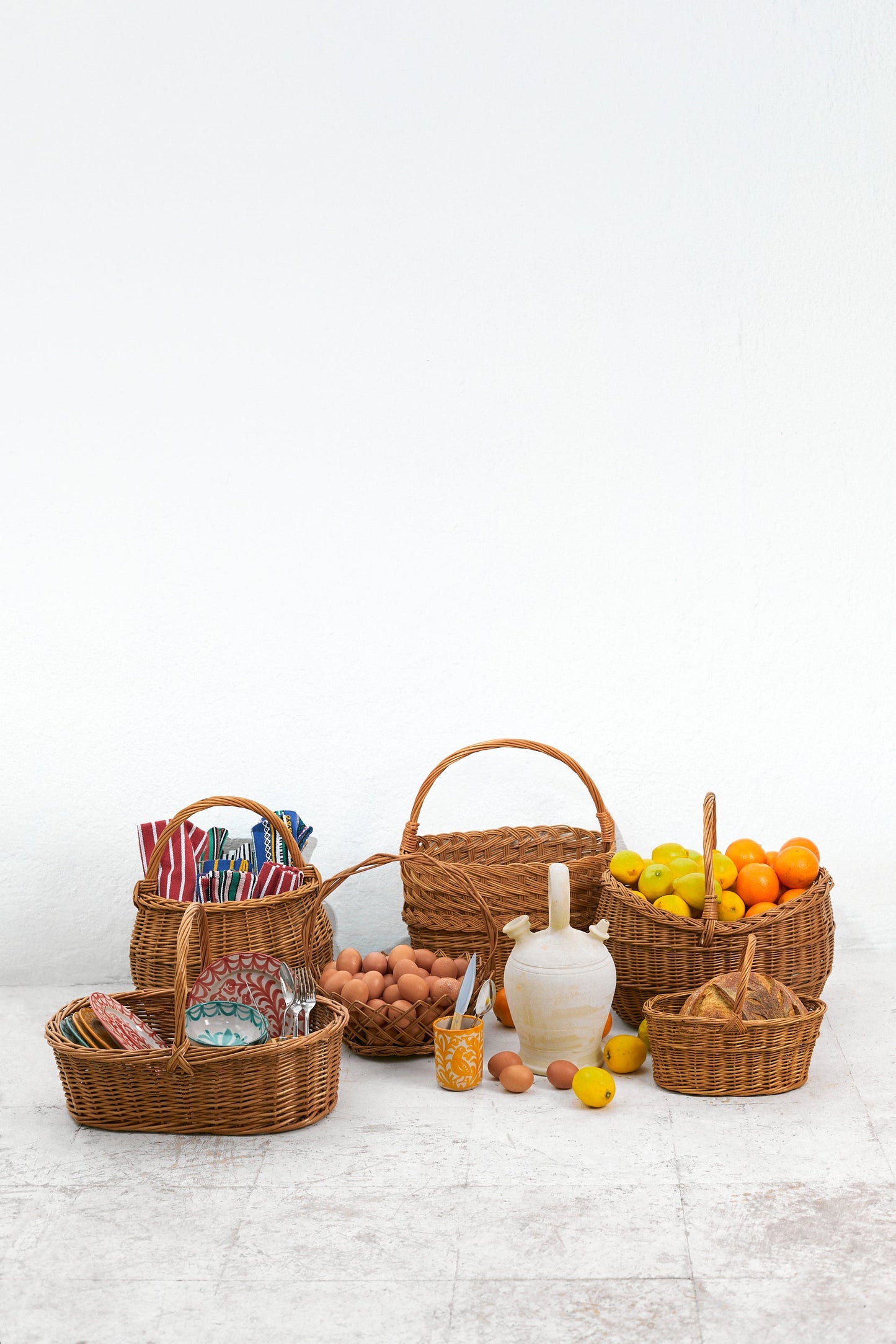 A collection of natural oval wicker baskets filled with fruit, eggs, and tableware against a white background.