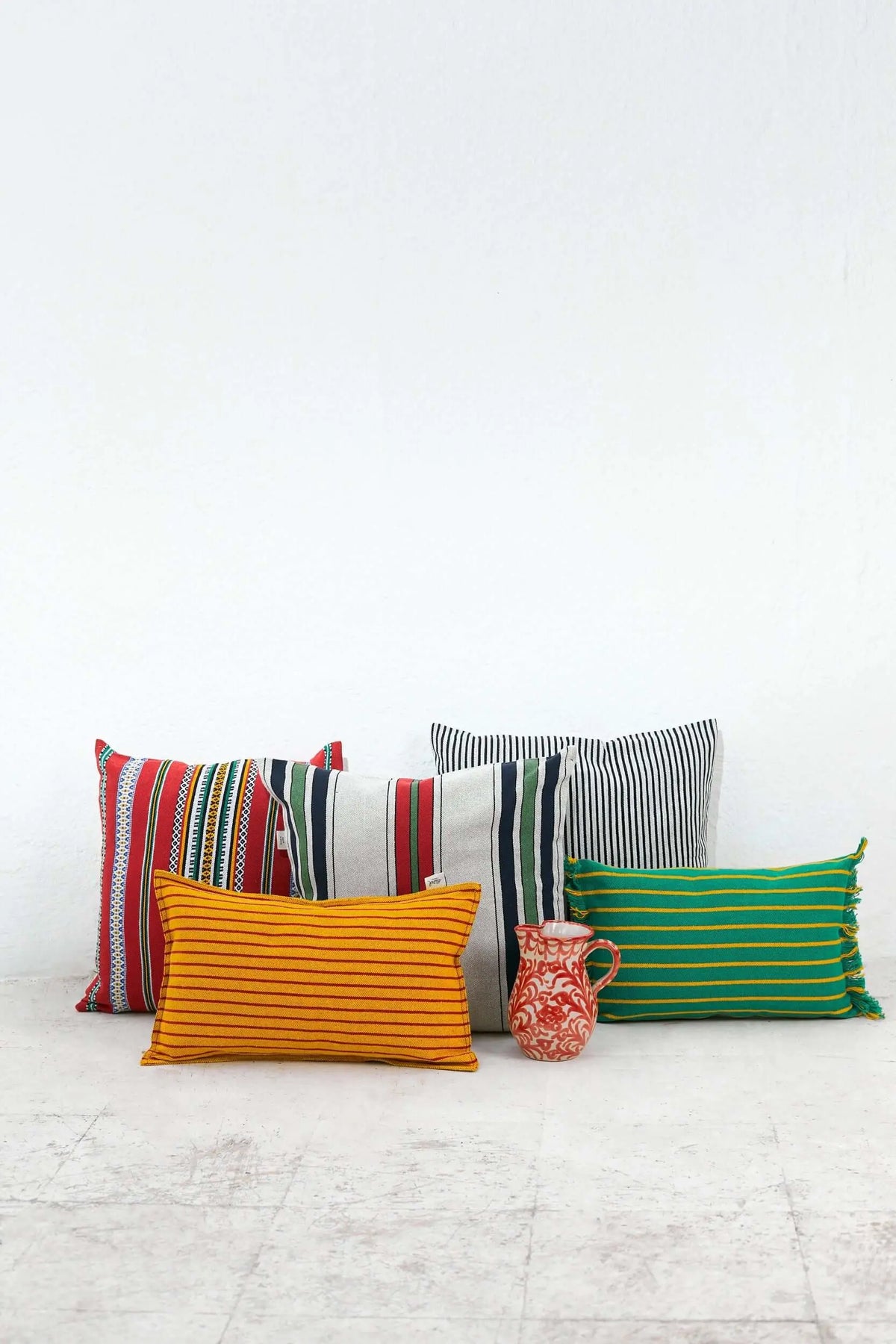 Cojines de loneta cuadrados y rectangulares con rayas multicolores, texturas textiles y una jarra de cerámica pequeña.