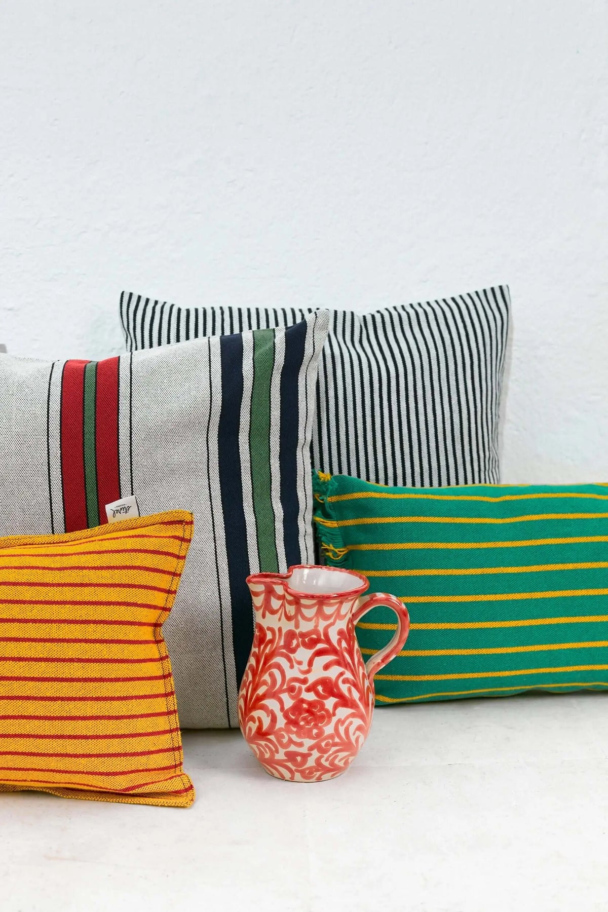 Conjunto de cojines cuadrados y rectangulares de loneta con estampados de rayas multicolores y una jarra de cerámica.