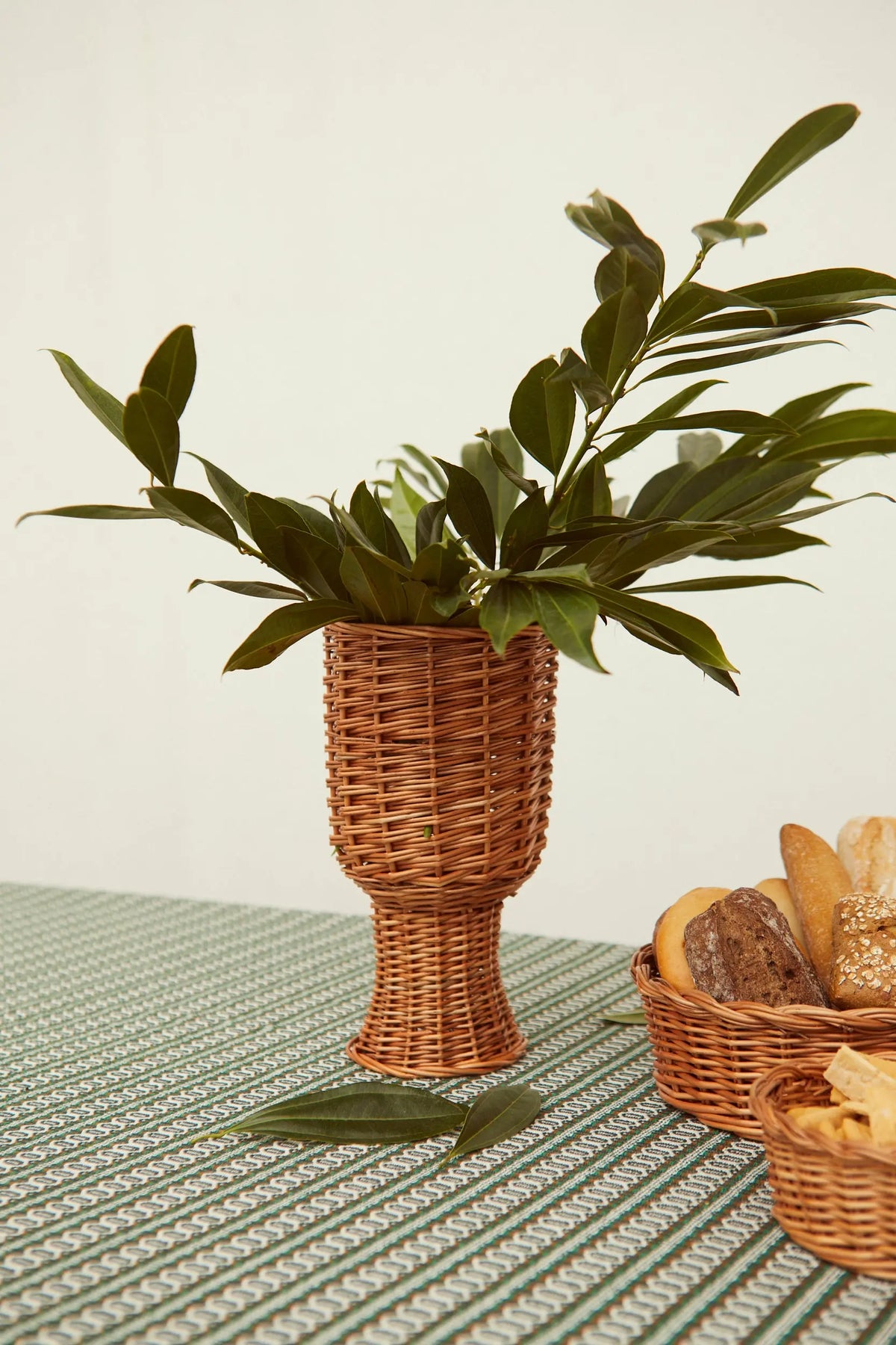 Jarrón de mimbre natural con diseño tipo copa, decorado con follaje verde sobre un mantel a rayas verdes y blancas.