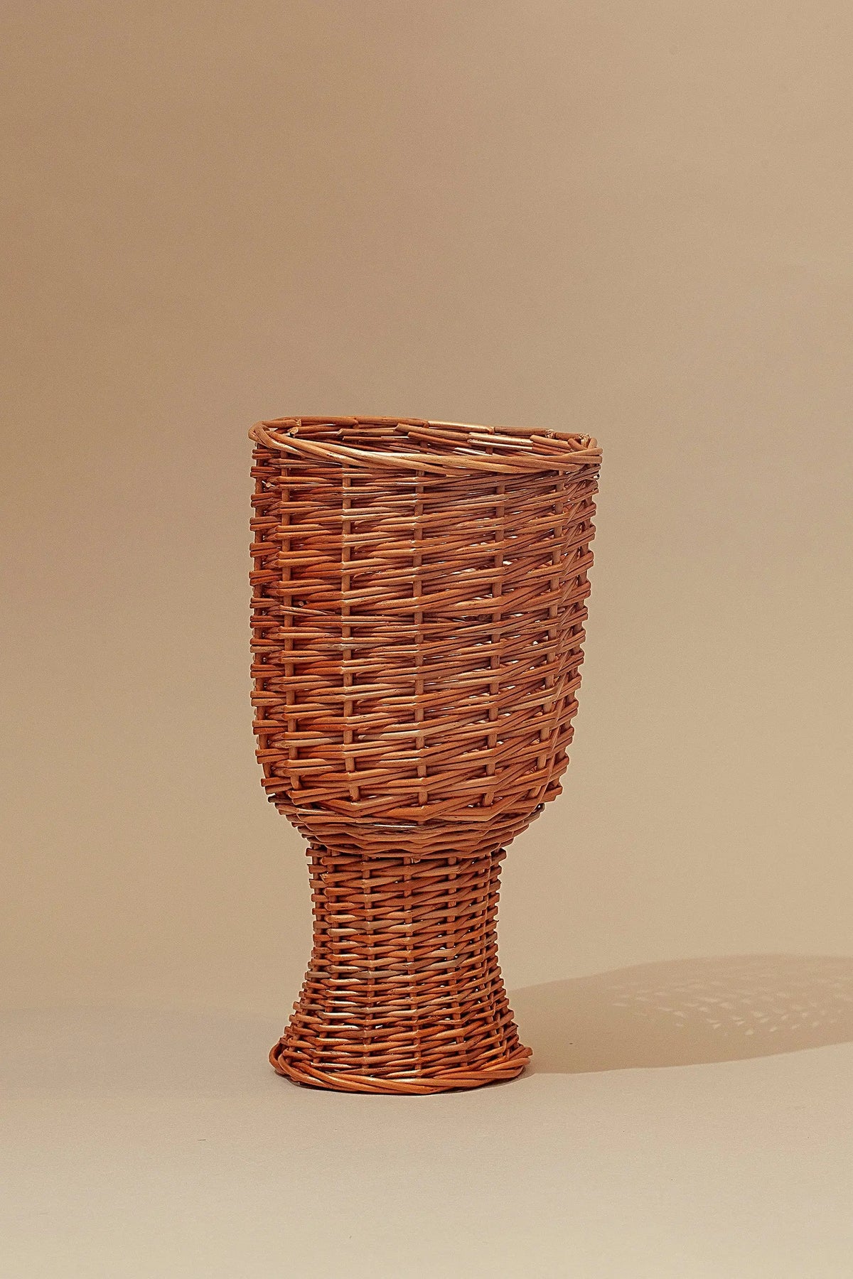 Jarrón pequeño de mimbre natural trenzado, color marrón claro, con forma de copa sobre fondo beige neutro.