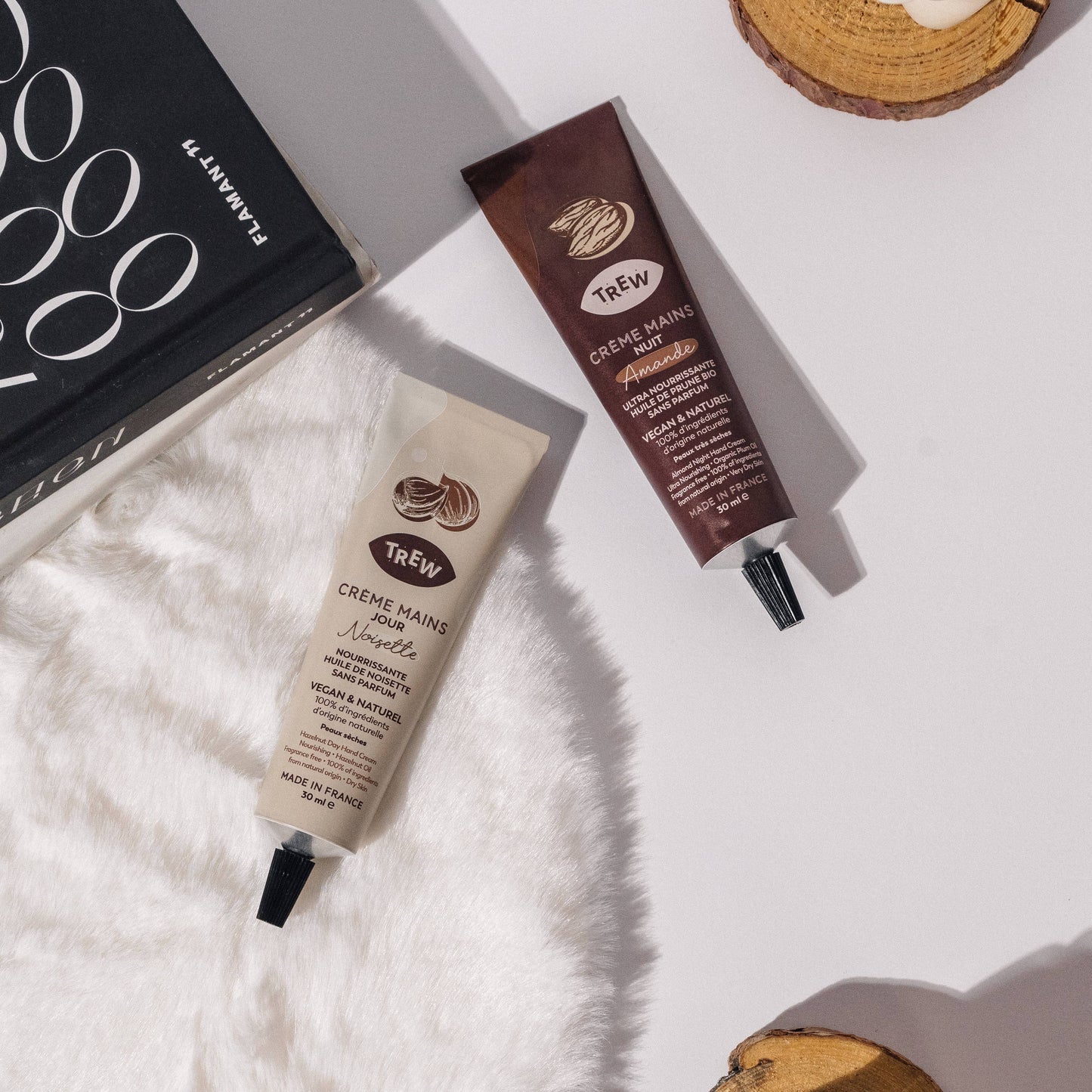 Two Trev hand cream tubes (beige and brown) on white fur, next to a black book and wood slices on a white background.