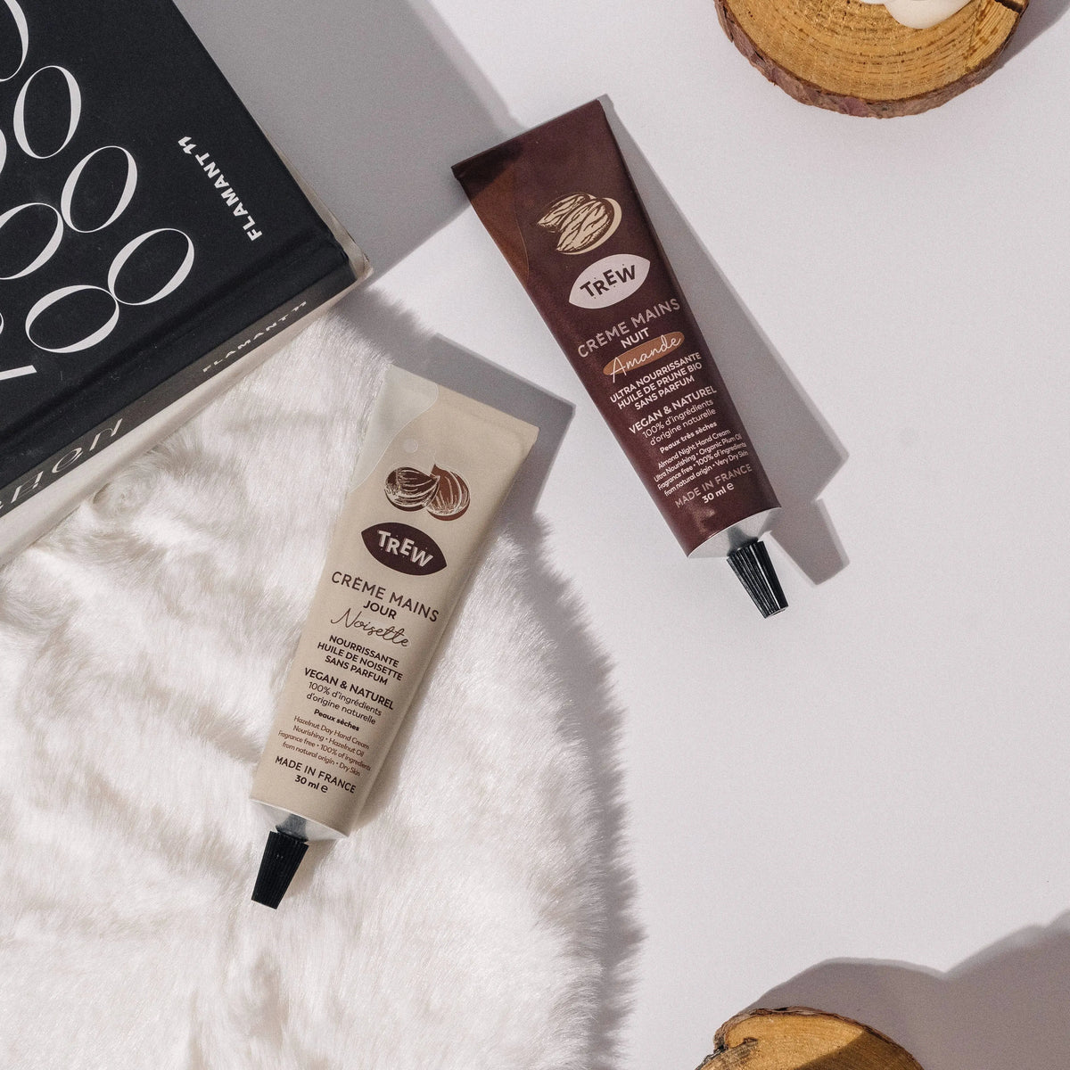 Two Trev hand cream tubes (beige and brown) on white fur, next to a black book and wood slices on a white background.