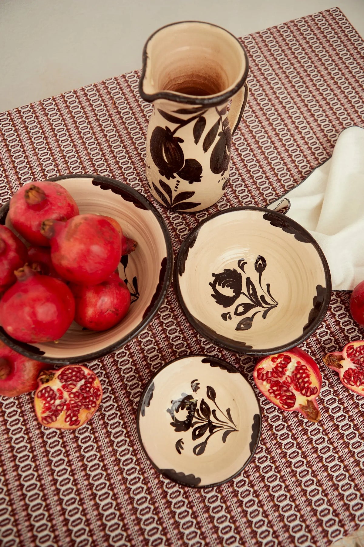 Set de cuencos de cerámica artesanal con motivos florales negros sobre fondo crema, rodeados de granadas frescas.