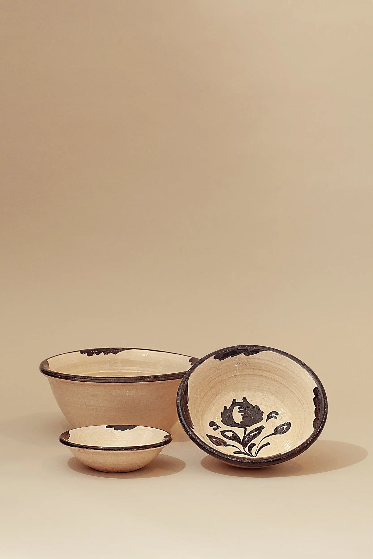 Three cream-colored ceramic bowls with hand-painted black floral patterns and dark trim against a beige background.