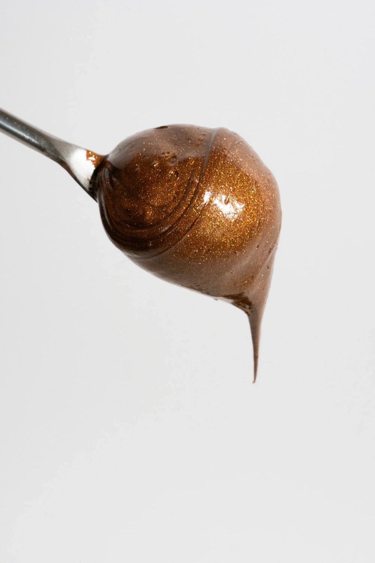 A dollop of shimmering, chestnut-brown lip balm on a metal spoon against a plain white background.