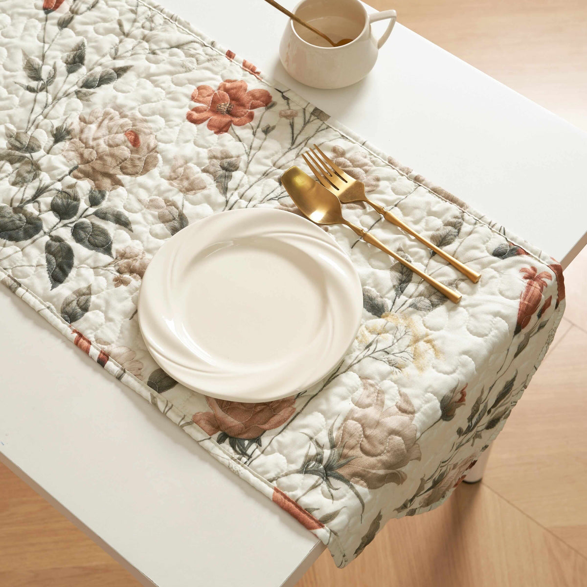 Quilted beige table runner with rose and olive floral print, featuring a white plate and gold cutlery set.
