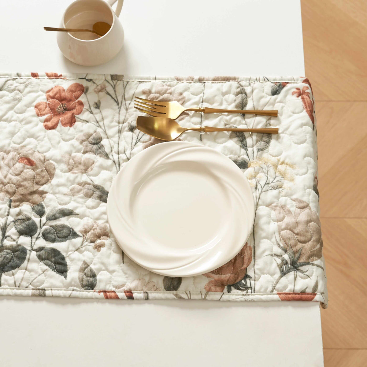 Quilted, rectangular table runner with floral pattern in beige, rose, and sage green, featuring a plate and cutlery.