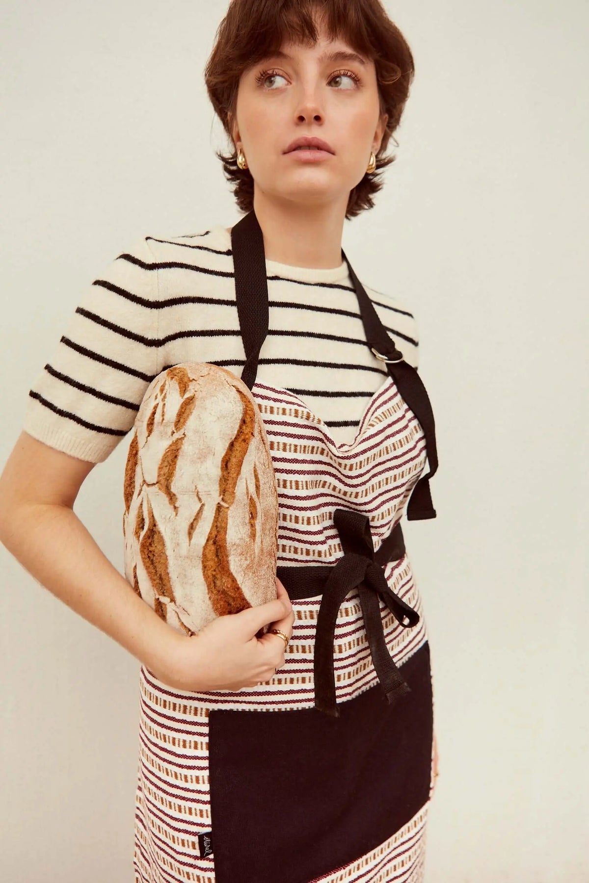 Mujer con delantal de tejido a rayas granates, lazo negro y bolsillo, sosteniendo un pan artesanal de masa madre.
