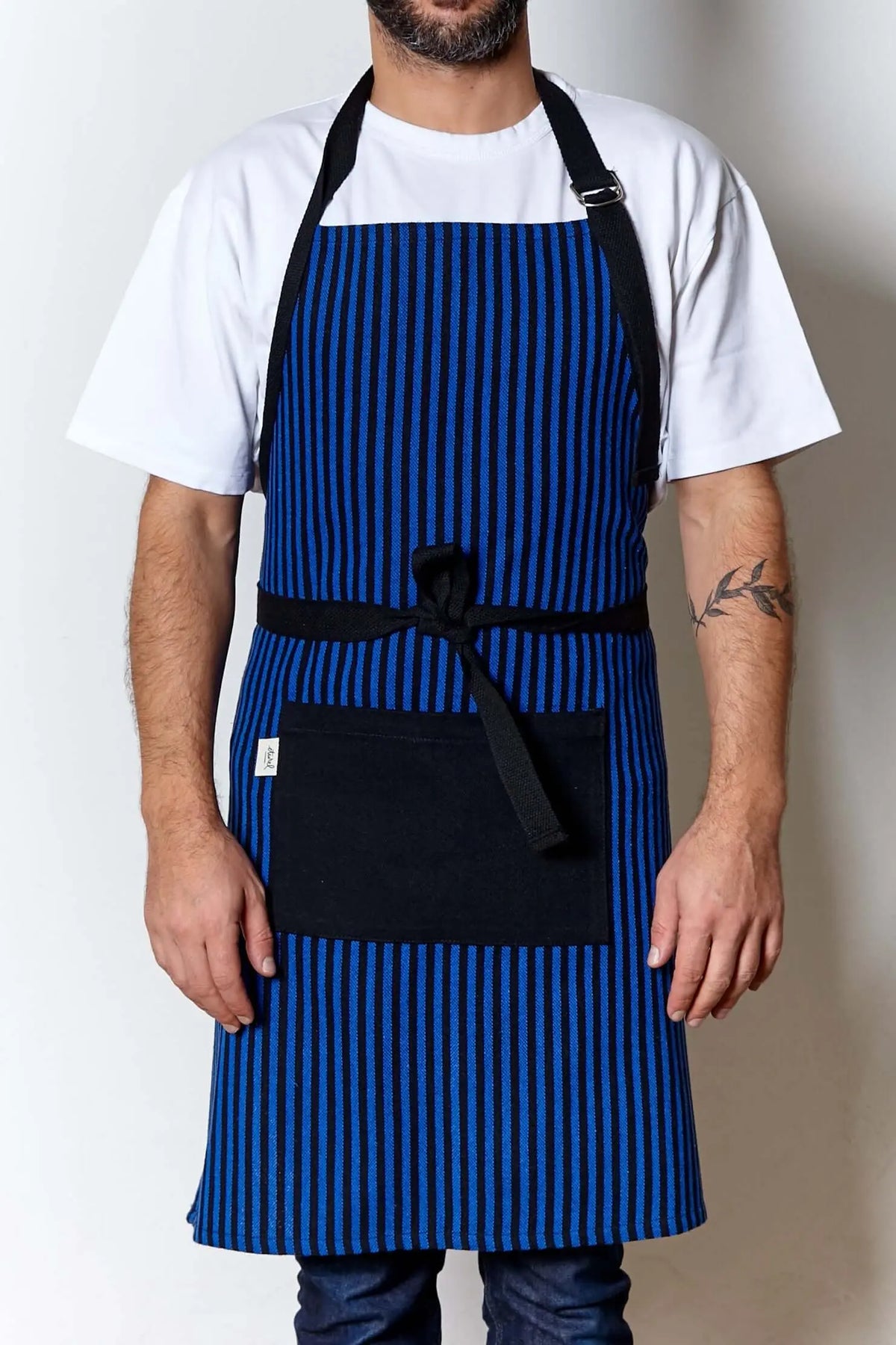 Delantal de loneta azul a rayas negras, con cintas y bolsillo central negro. Incluye hebilla ajustable en el cuello.