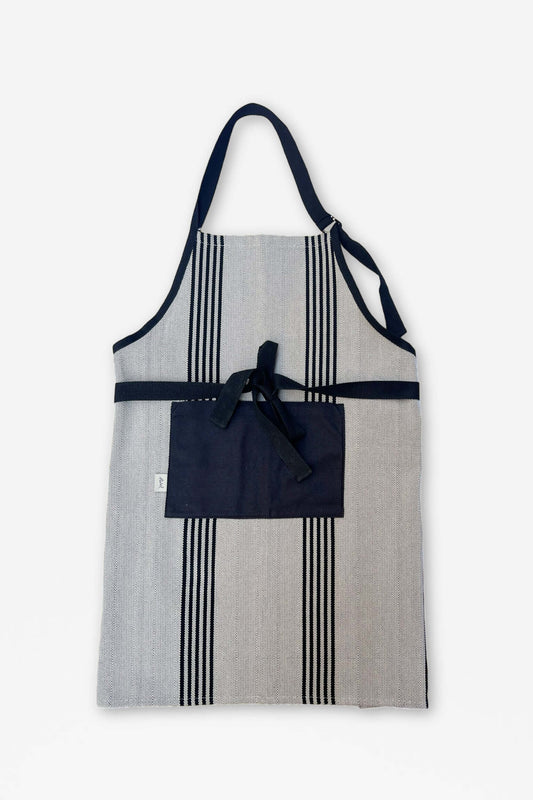 Delantal de sarga gris con rayas verticales negras, bolsillo central, tiras negras y acabado estilo rústico.